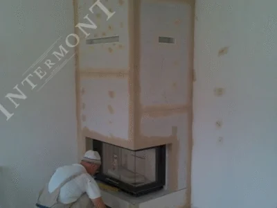 Obloge kamina - Intermont Majstor u radnoj odjeći čuči ispred kamina u izgradnji. Obloga je od gips-vlaknastih ili mineralnih vatrootpornih ploča. Vidljivi su ventilacijski otvori za topli zrak na gornjem dijelu i radovi na podnožju kamina.