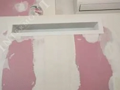 Obloge kamina - Intermont Uski, dugački, bijeli pravokutni otvor za zrak u glatkoj, bijelo obojenoj površini zida. Dizajn otvora je minimalistički, bez vidljive rešetke, integriran u oblogu kamina u fazi izgradnje/završne obrade.